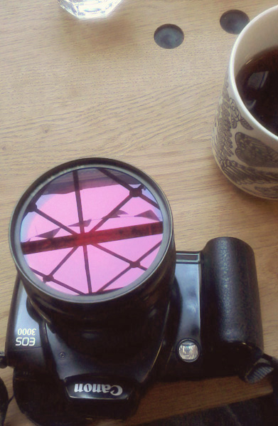 Spiegelung einer Wimpelkette an einer x-verstrebten Eisenkonstruktion im Rotfilter eines Kameraobjektivs, auf einem Kaffeetisch liegend. Tisch und Kaffeetasse puzzeln sich zu einem Gesicht.